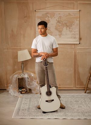 A person in a white t-shirt sits cross-legged on a patterned rug with an acoustic guitar in a minimalist room.