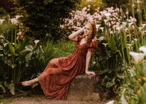 A person in a flowing red dress reclines on a stone wall surrounded by blooming calla lilies.