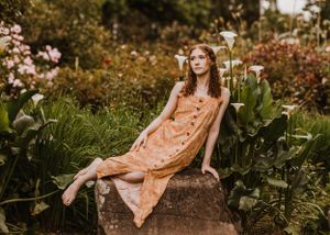 A person in a peach dress sits on a rock surrounded by flower gardens on a sunny day.