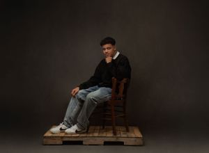 Moody portrait of person seated on wooden chair against dark background.
