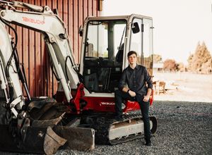 A construction worker poses with a Takeuchi mini excavator on a gravel surface.