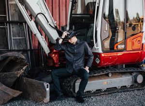 A person sits on construction equipment wearing dark clothing and a baseball cap.