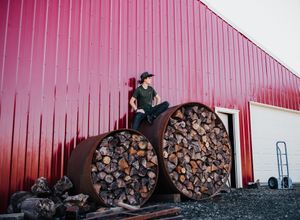 A person sits atop large circular wood storage containers against a red metal building.