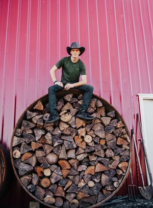 A cowboy-style portrait on stacked firewood against a red metal wall.