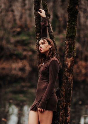 A moody autumn forest portrait of someone in a brown dress leaning against a tree trunk near water.