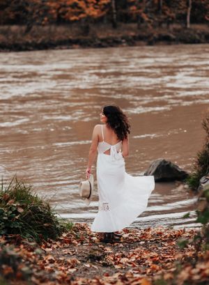 Flowing white dress creates movement along riverbank covered in fall leaves.