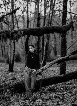 A black and white forest portrait of someone in jeans leaning against a fallen tree.