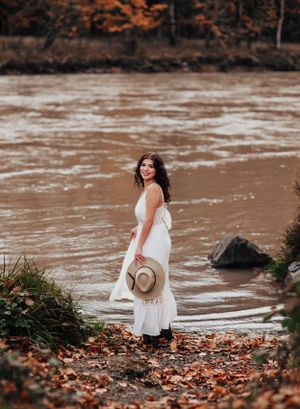 White dress and hat ensemble photographed by muddy river with autumn foliage.