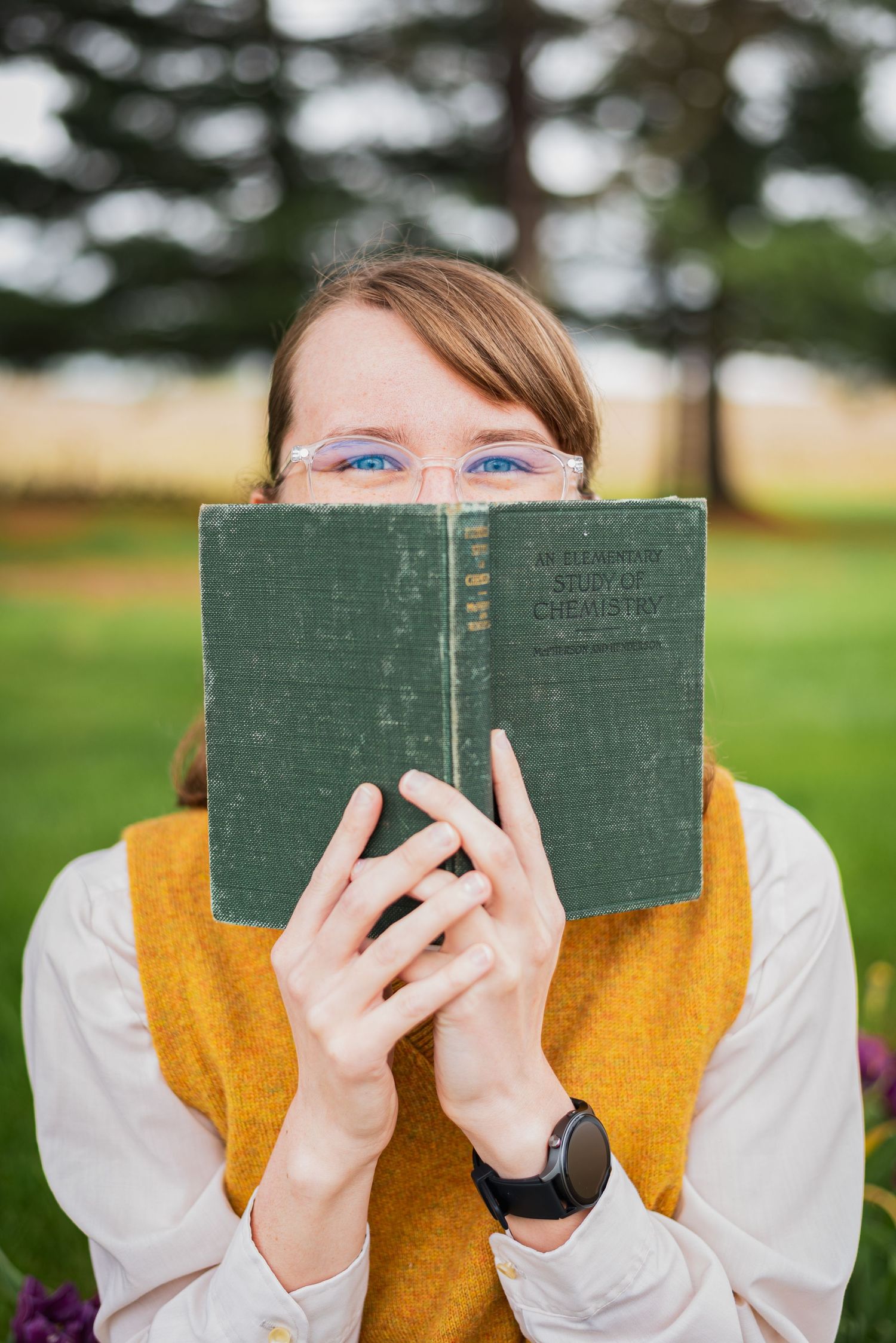 Person in yellow vest holding vintage book covering face outdoors.
