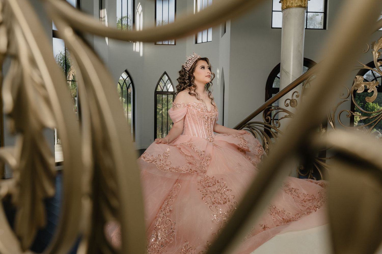 A person in a blush pink ballgown poses on an elegant staircase framed by golden architectural elements.