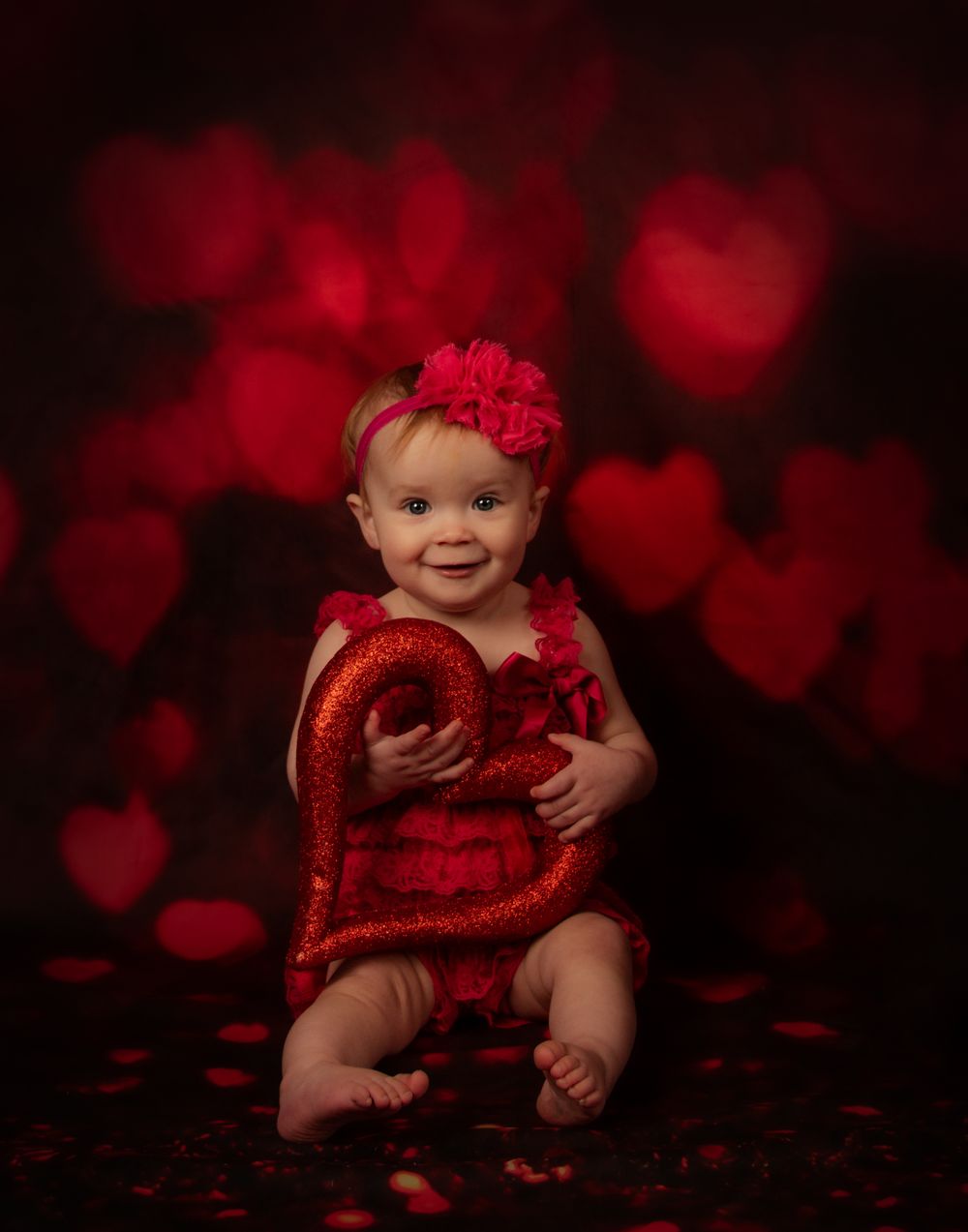 Baby in red tutu dress photographed against dark background with heart-shaped bokeh lights.