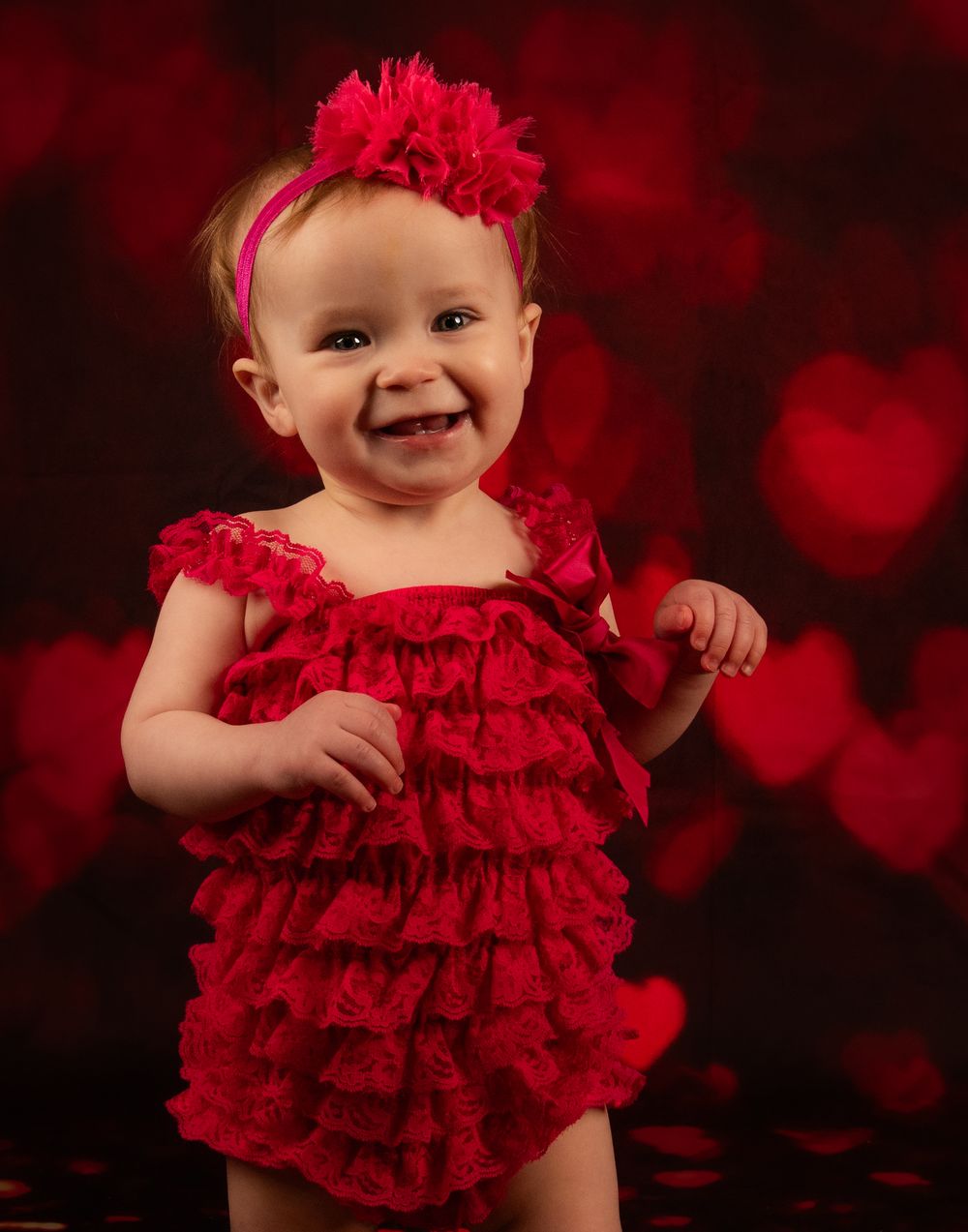 Baby in red ruffled dress poses against dark red background with bokeh hearts effect.