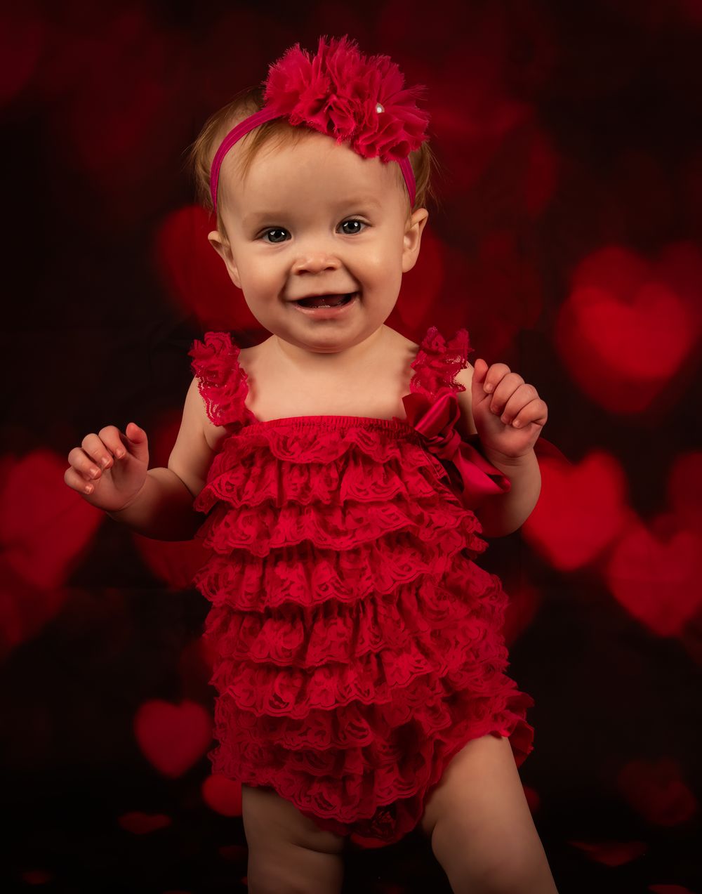 A joyful baby wearing a red ruffled romper and matching headband against a dark background with heart bokeh.