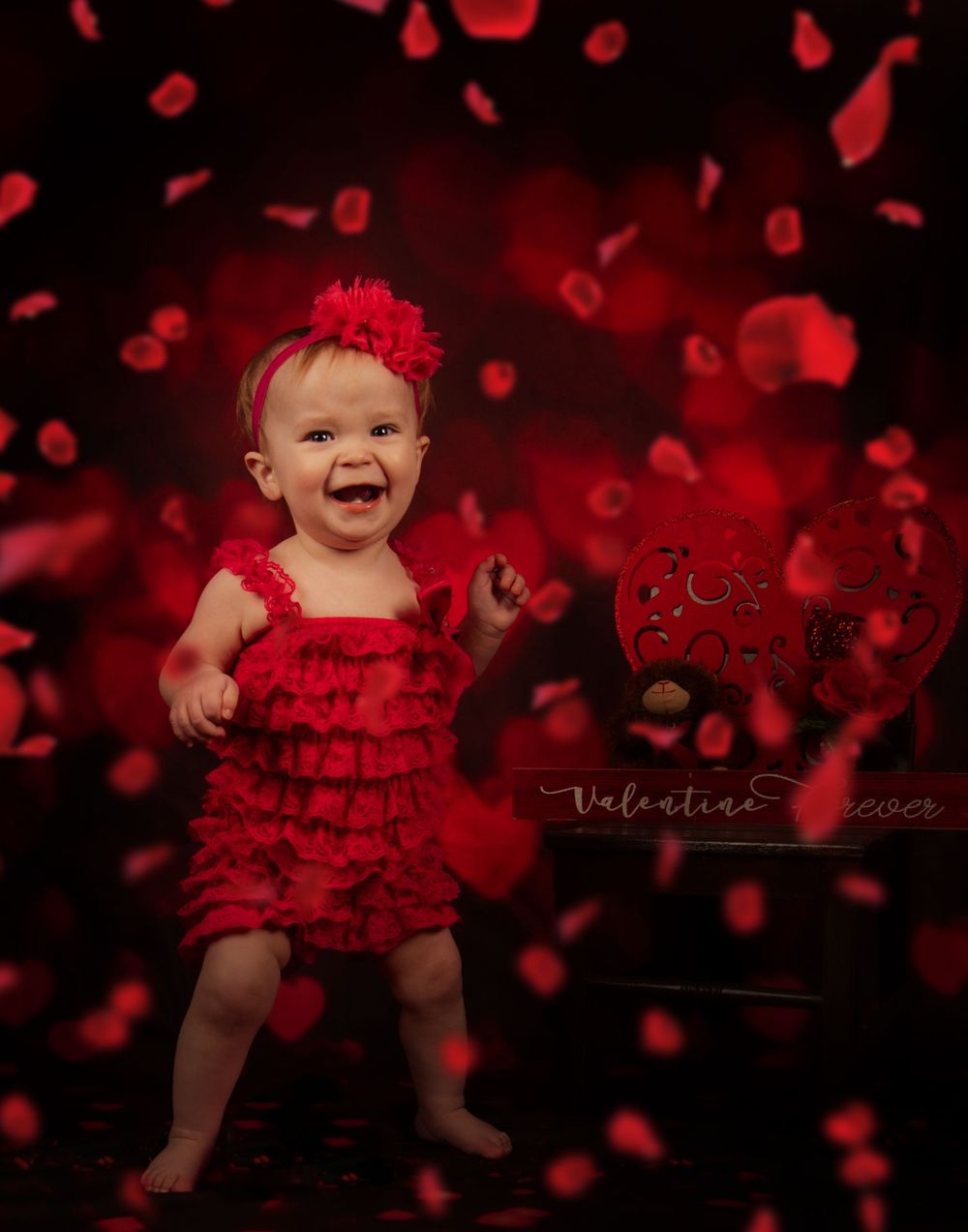 Baby in red ruffled dress poses against dark background with floating red rose petals for Valentine's Day photo shoot.