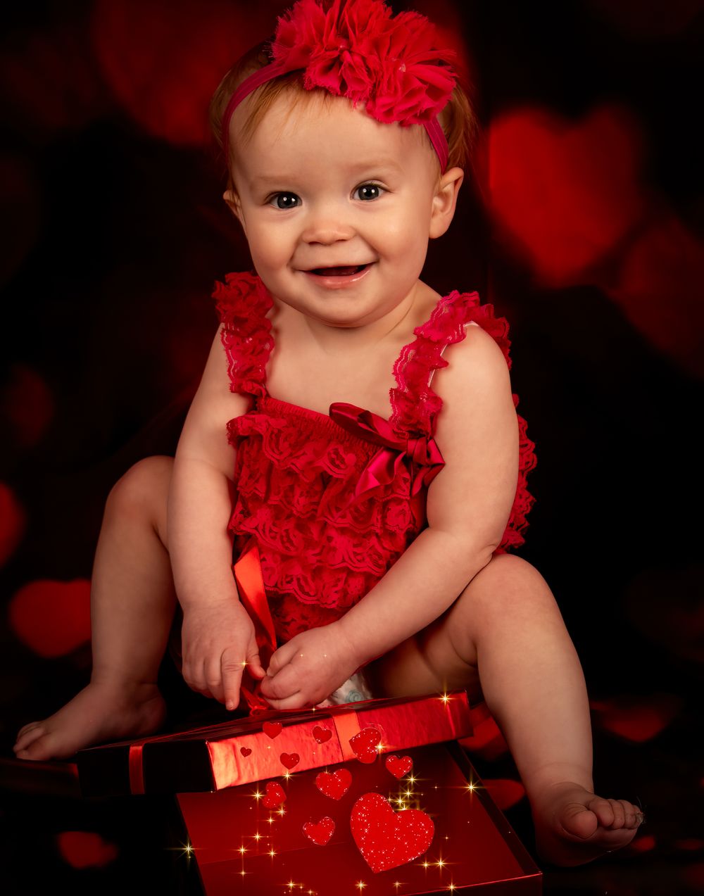 Baby in red ruffled dress and matching bow poses against dark background with red hearts and sparkly lights.