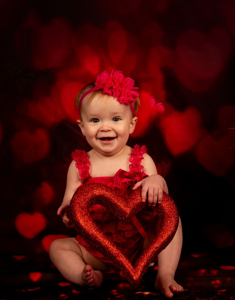 A baby in red attire sits with a sparkly red heart decoration against a dark background with heart-shaped bokeh effects.