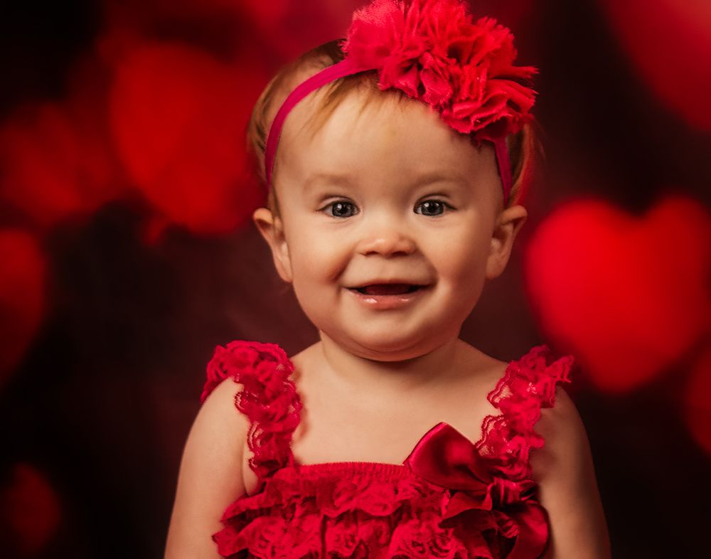 Baby wearing red flower headband and dress smiles against dark background with red bokeh lighting effect.
