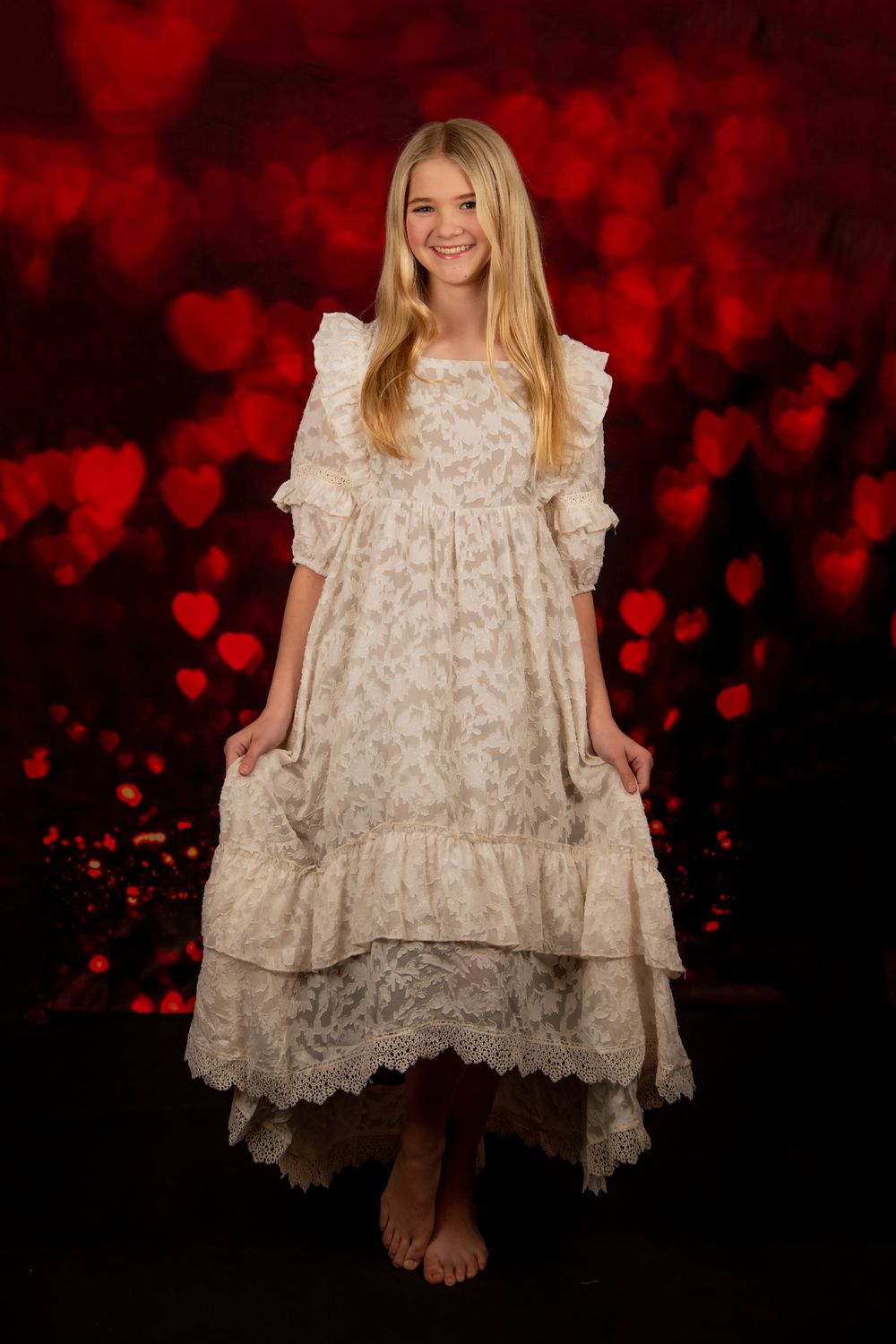 A young girl in a vintage-style cream lace dress poses against a dark red bokeh background.