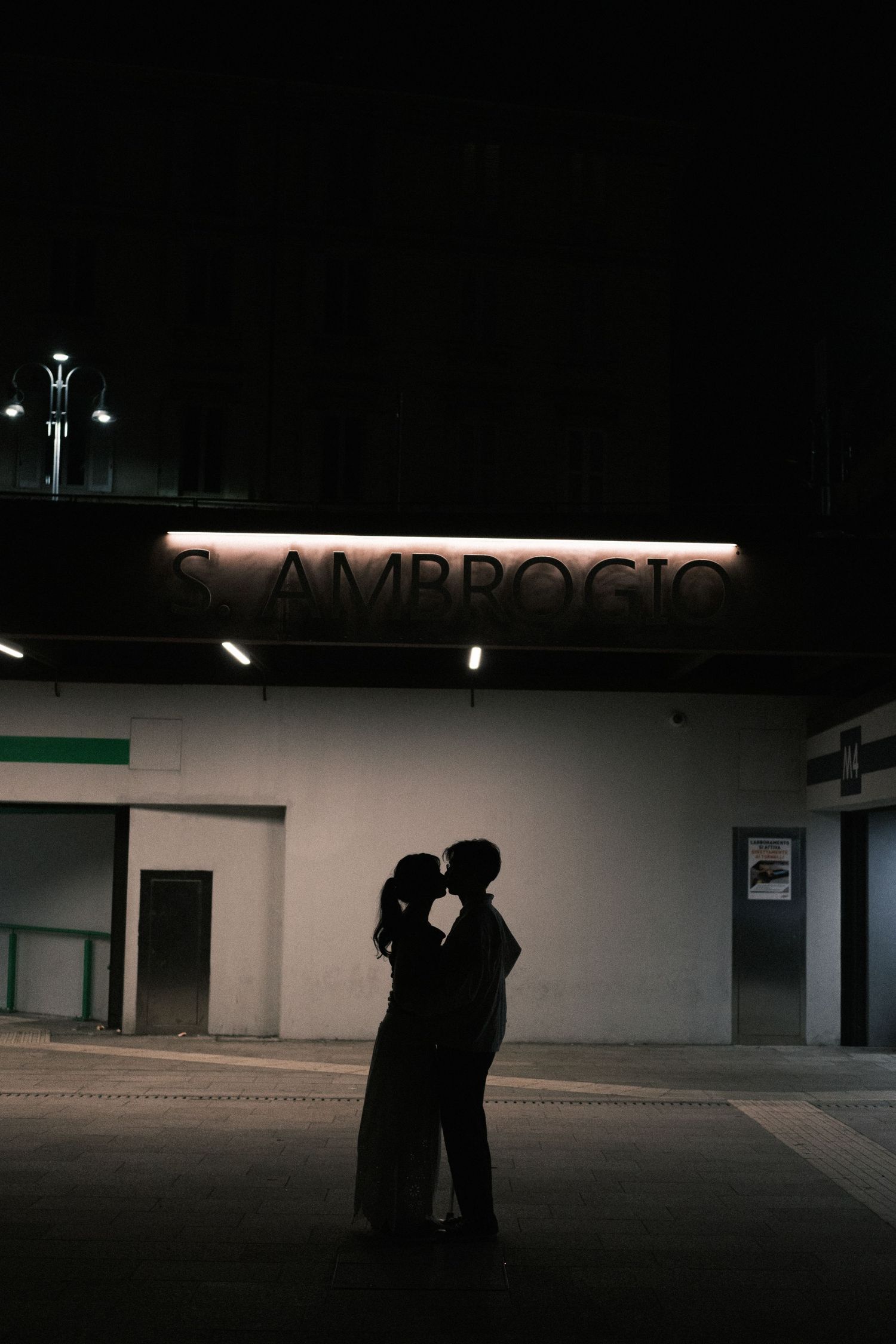 A dark silhouette of two figures standing in a dimly lit parking lot at night with illuminated signage in the background.