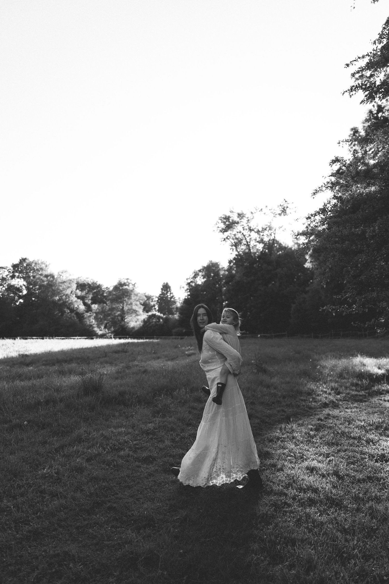 Black and white photo of a bride in a lace gown holding a child in a sunlit grassy park surrounded by trees.