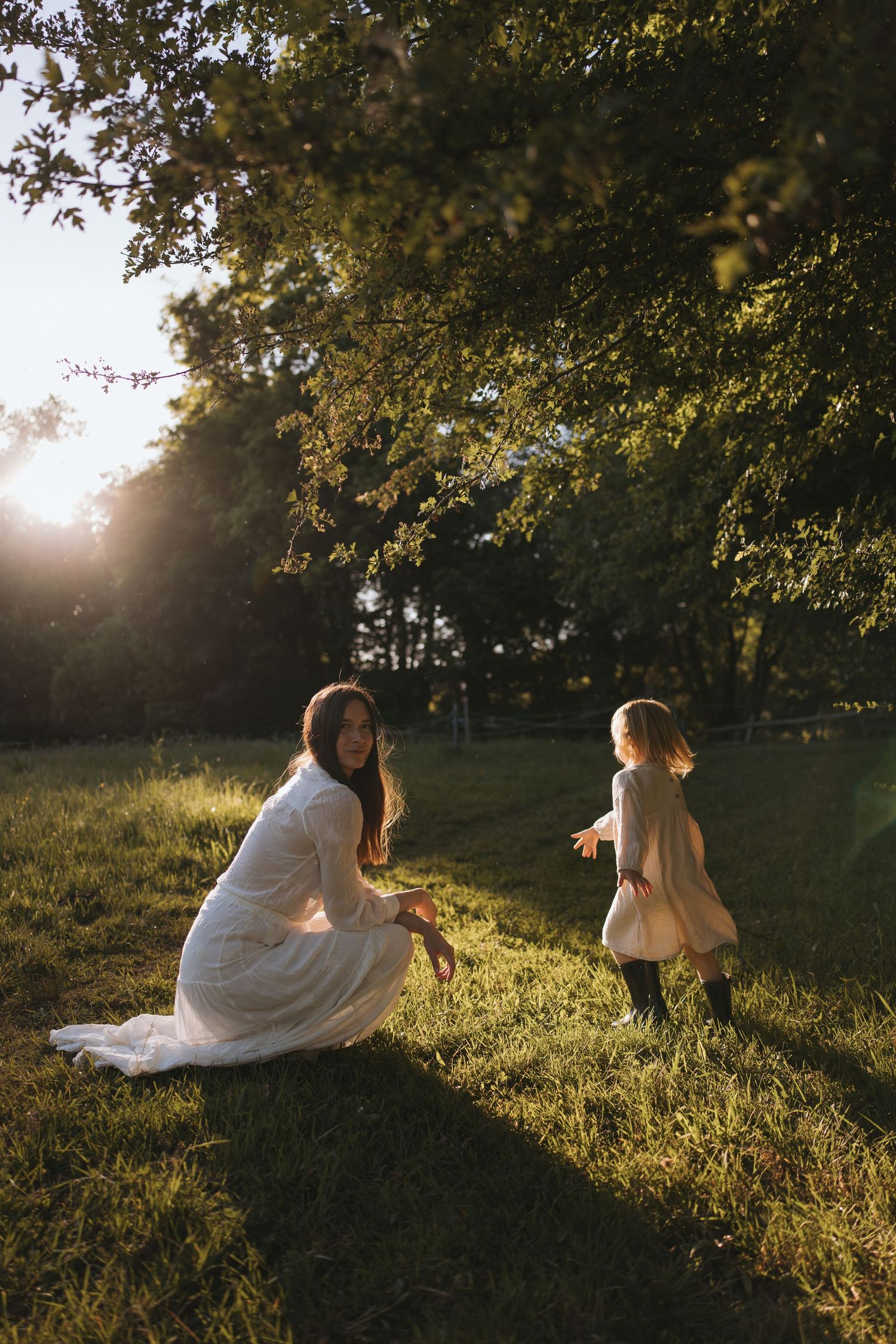 A mother in a white dress crouches on green grass as her young daughter runs toward her in golden sunset light.