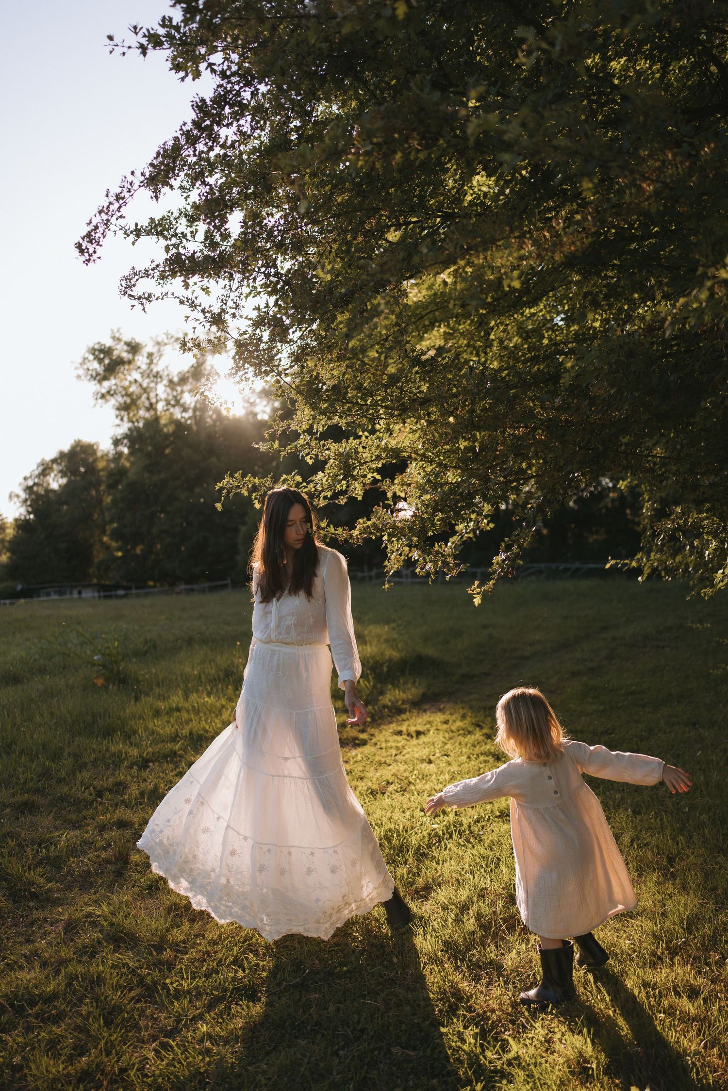 A mother and daughter in matching white dresses dance together in a sunlit green park near a large tree.