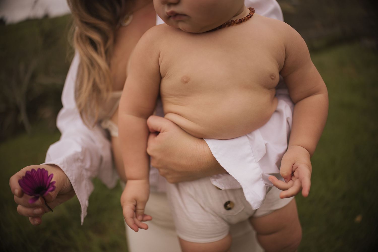 A mother holding her chubby baby outdoors, with a pink flower in her hand, both dressed in white.