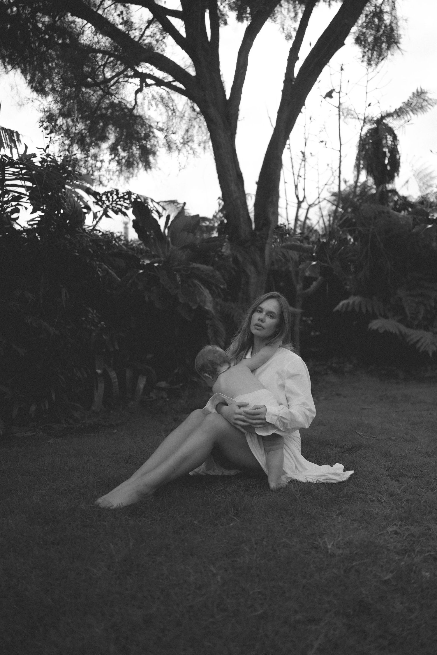 Black and white photo of a woman in a white robe sitting on grass in a lush garden with tropical trees.