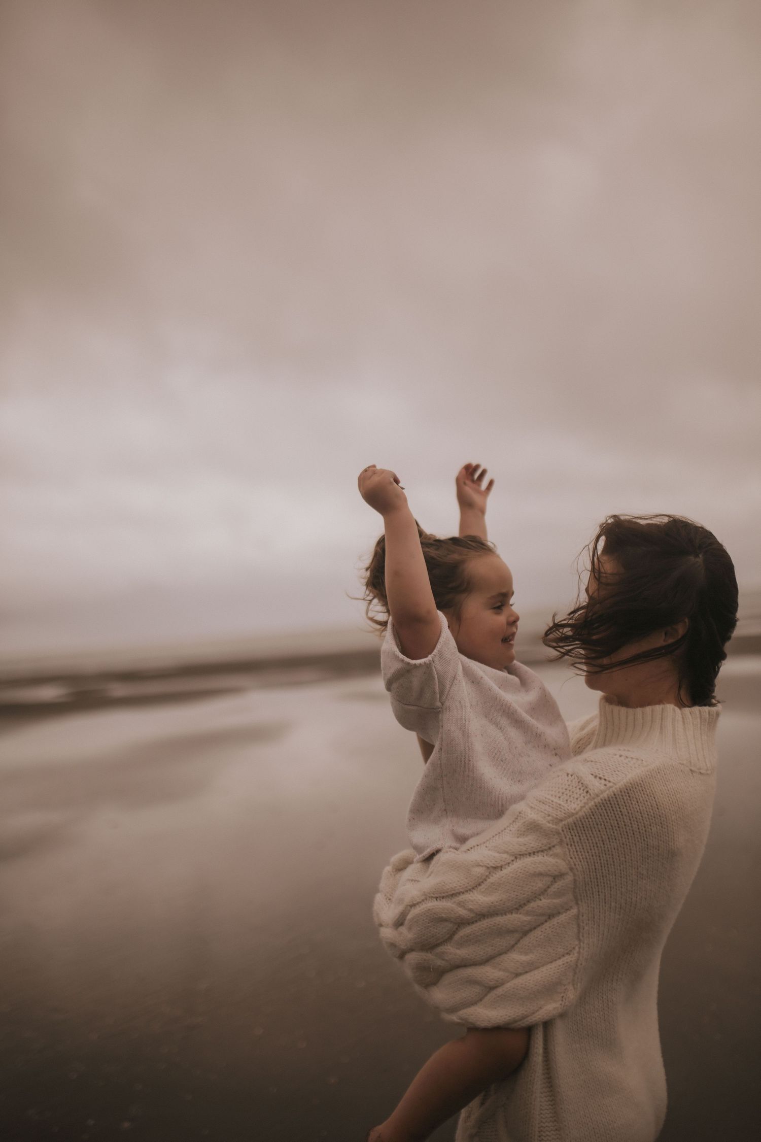 A mother in a white cable-knit sweater holds her toddler on a misty beach, as the child joyfully raises their arms in New Zealand.