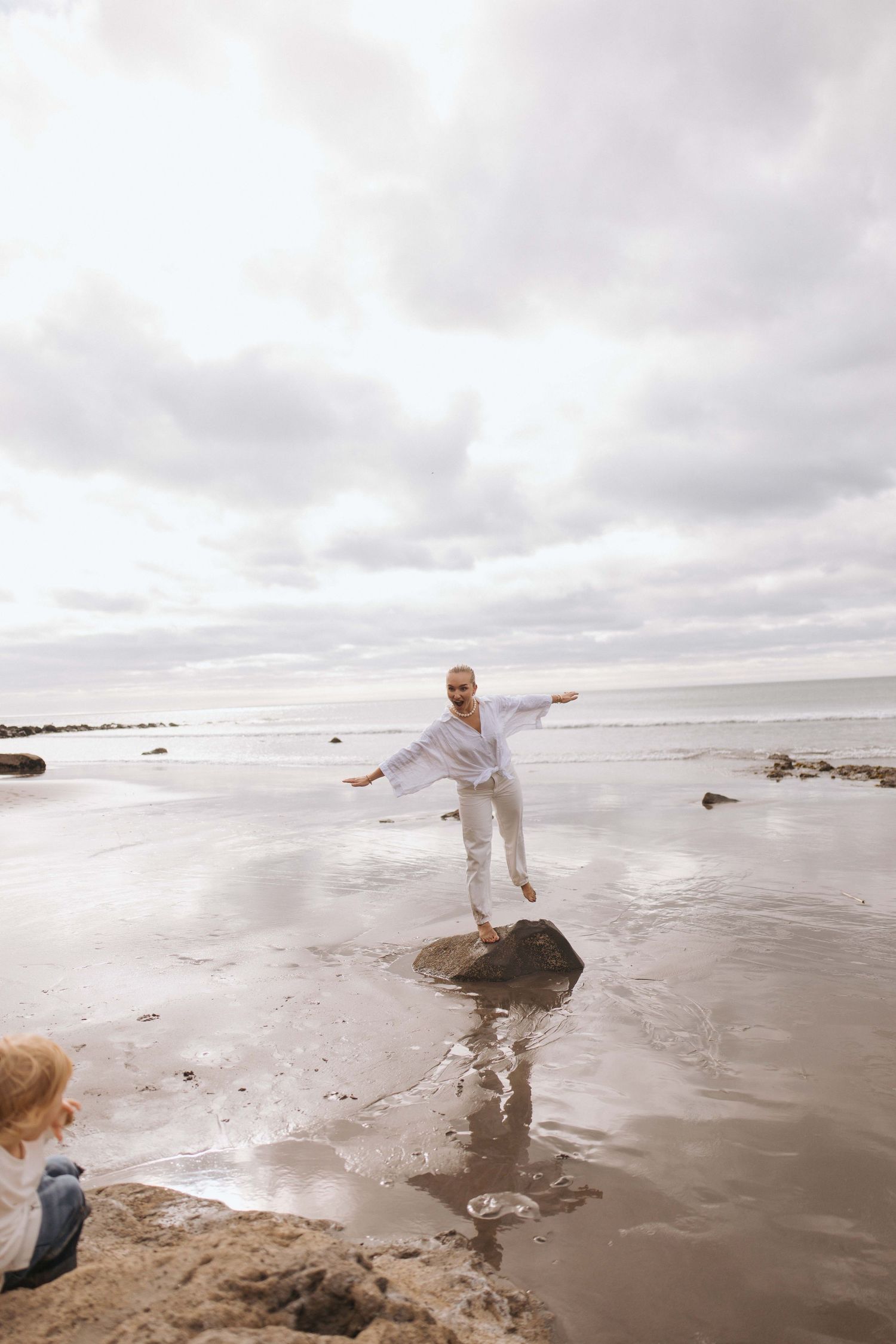 Person balancing on a rock at the beach with arms outstretched, wearing white outfit on a cloudy day.