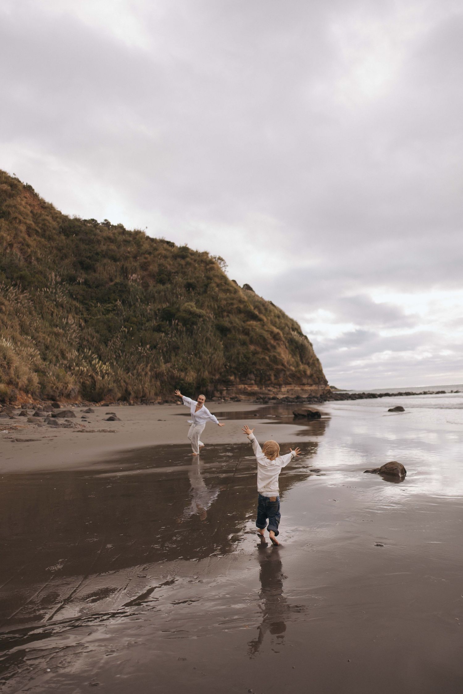 Two children playing joyfully on a moody, overcast beach with dramatic cliffs and golden foliage in the background.
