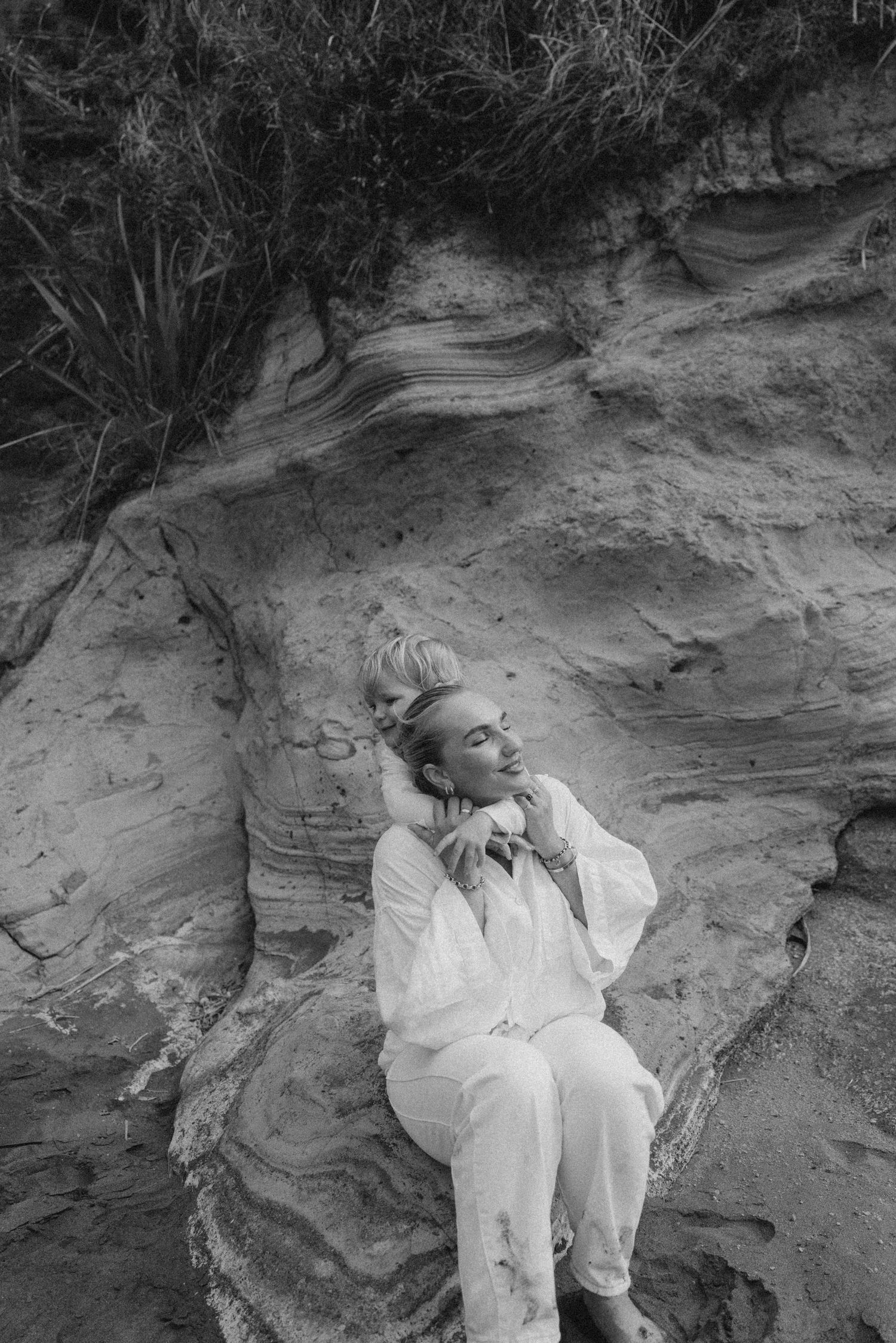 Black and white photo of a woman in white outfit resting against layered sandstone rock formation outdoors.