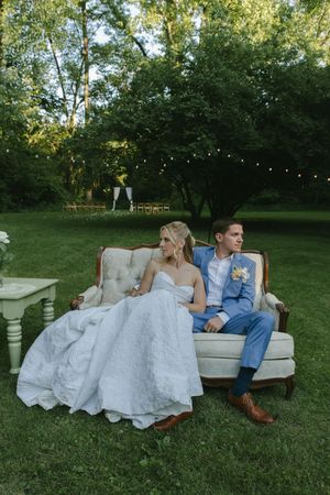 A bride in white and groom in blue sit together on an elegant outdoor sofa beneath string lights.
