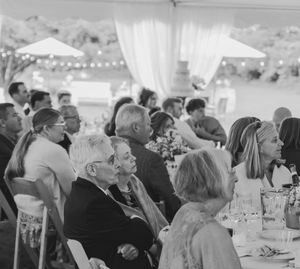 Wedding guests seated under a white reception tent with string lights and decorative details.