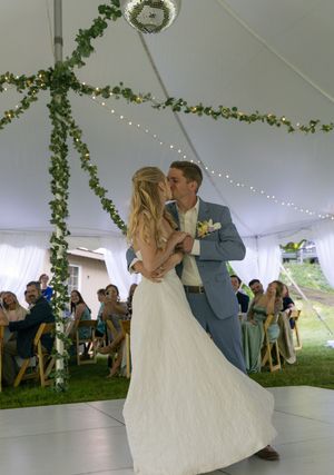 A couple shares their first dance under a white tent decorated with greenery and string lights.