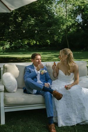 A couple in wedding attire share ice cream cones while sitting on an outdoor sofa during their garden reception.
