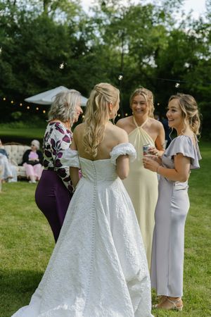 Bridesmaids in lavender dresses gathered around bride in white wedding gown on grass.