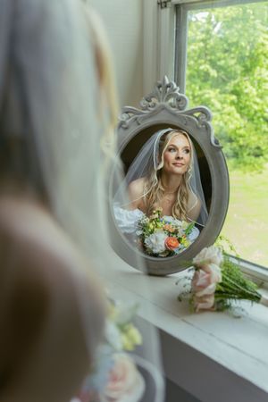 Vintage mirror reflects a bride with flowers gazing out a window on her wedding day.