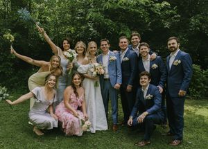 Wedding party members in blue suits and pastel dresses posing playfully on grass.