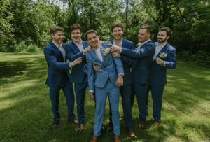 Groomsmen in blue suits pose playfully together on a sunny lawn.