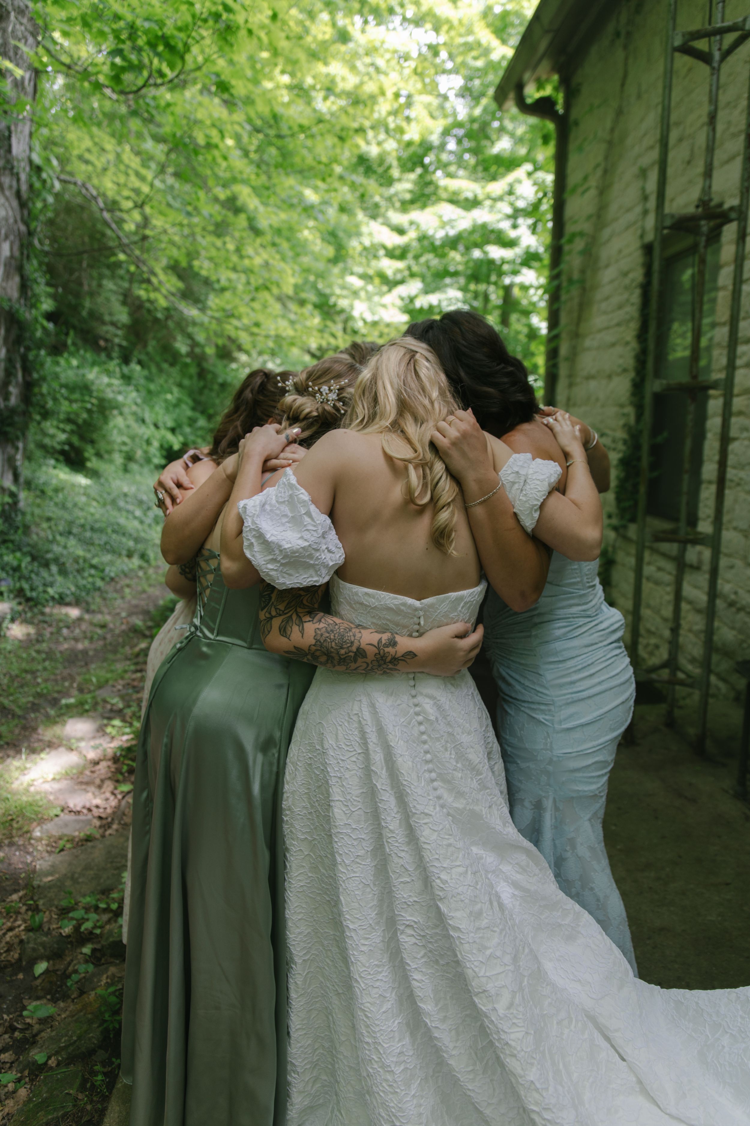 Bridesmaids in pastel dresses share a group embrace in a garden setting.