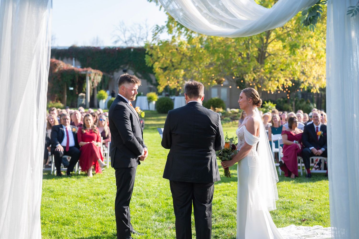 Outdoor wedding ceremony setup with white draped fabric and guests gathering on a sunny autumn day.
