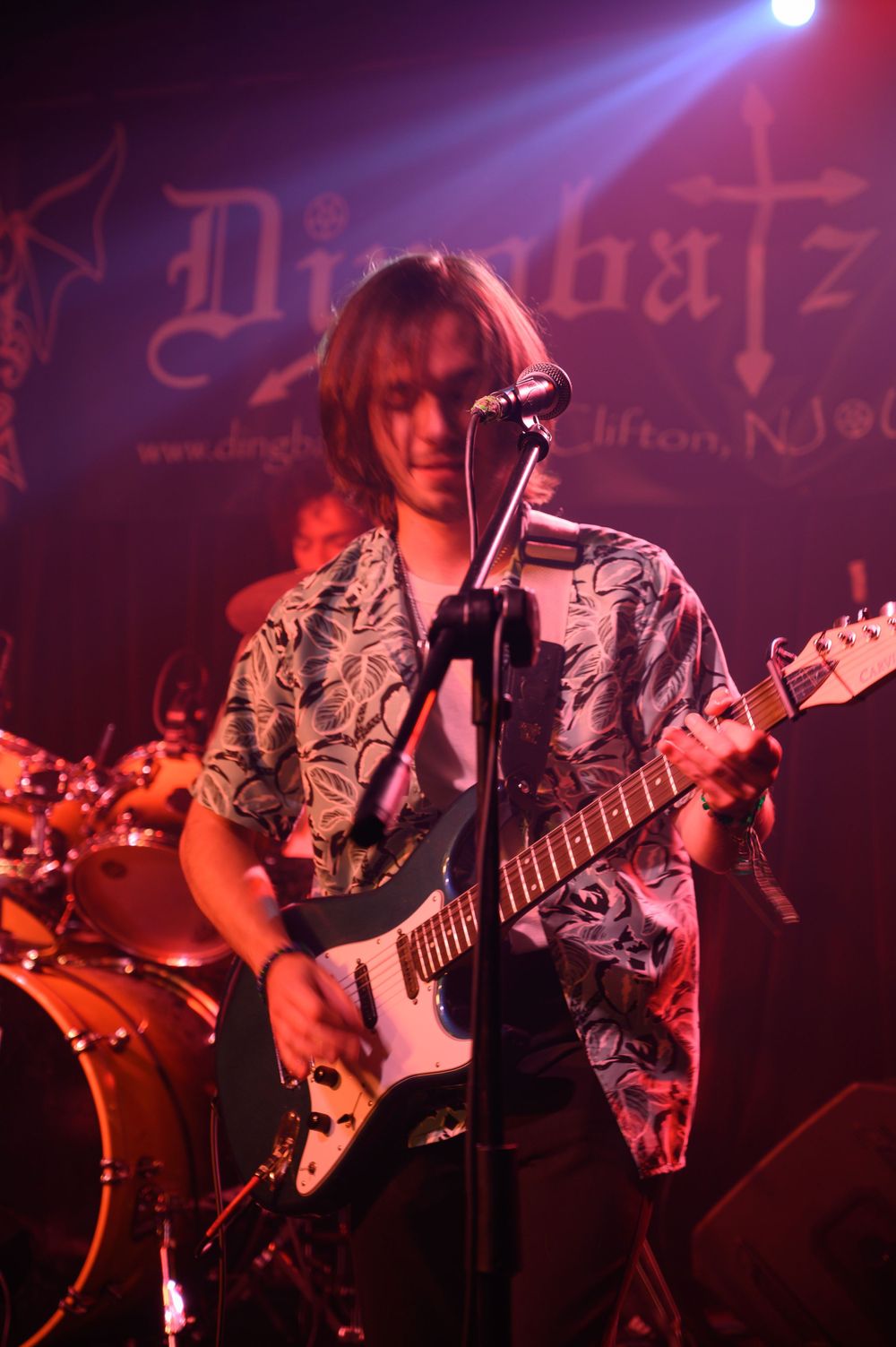 A guitarist performs on stage wearing a patterned shirt, playing an electric guitar under colorful stage lighting.