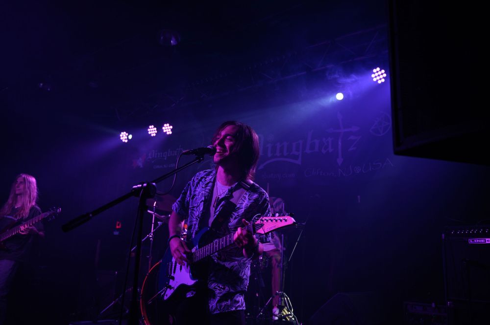 A musician performs on stage with a guitar under blue and purple stage lighting during a live concert performance.
