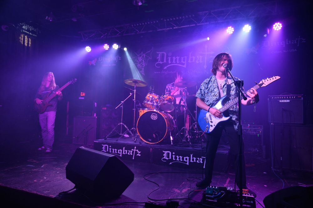 A rock band performs on stage with purple and blue lighting at what appears to be a live music venue.
