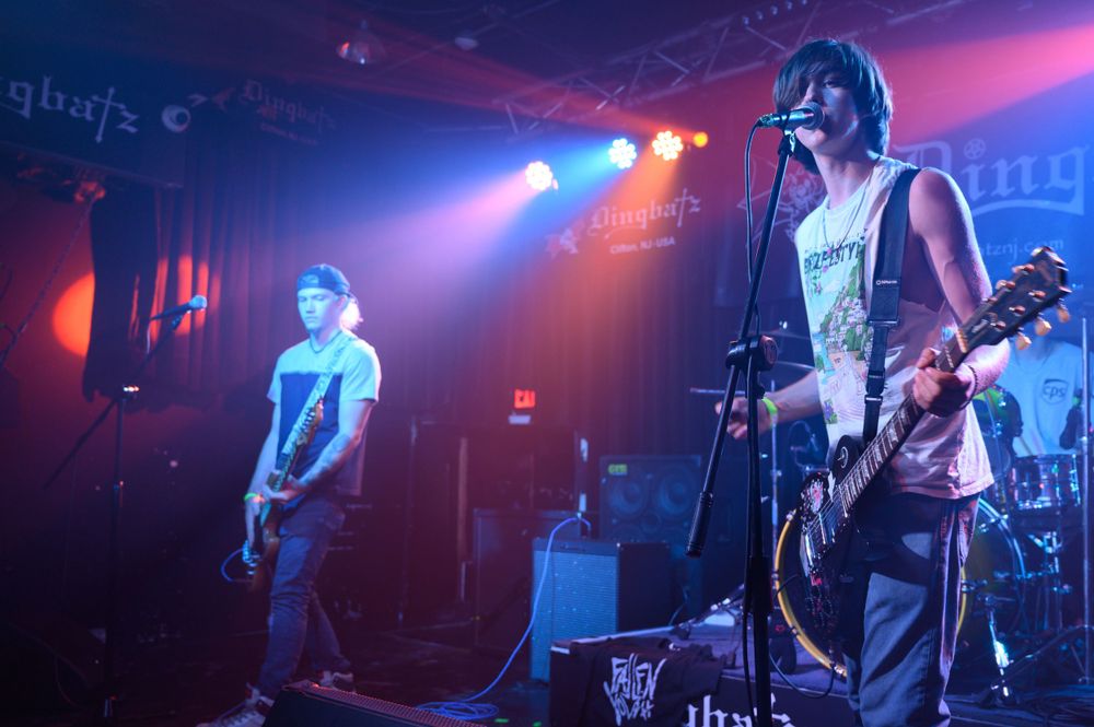 Musicians perform on stage at a rock concert with purple and blue lighting effects illuminating the dark venue.