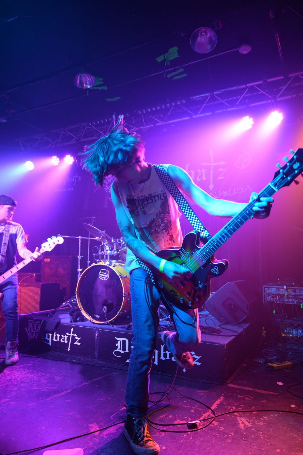 Musician plays electric guitar on a purple-lit concert stage with dramatic lighting and stage equipment visible.