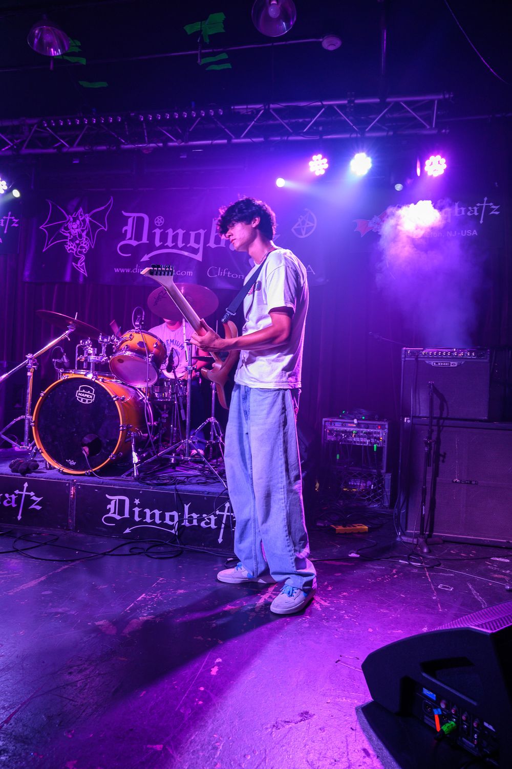 Musician performs on stage bathed in purple lighting at Dinghof venue with drum kit visible in background.