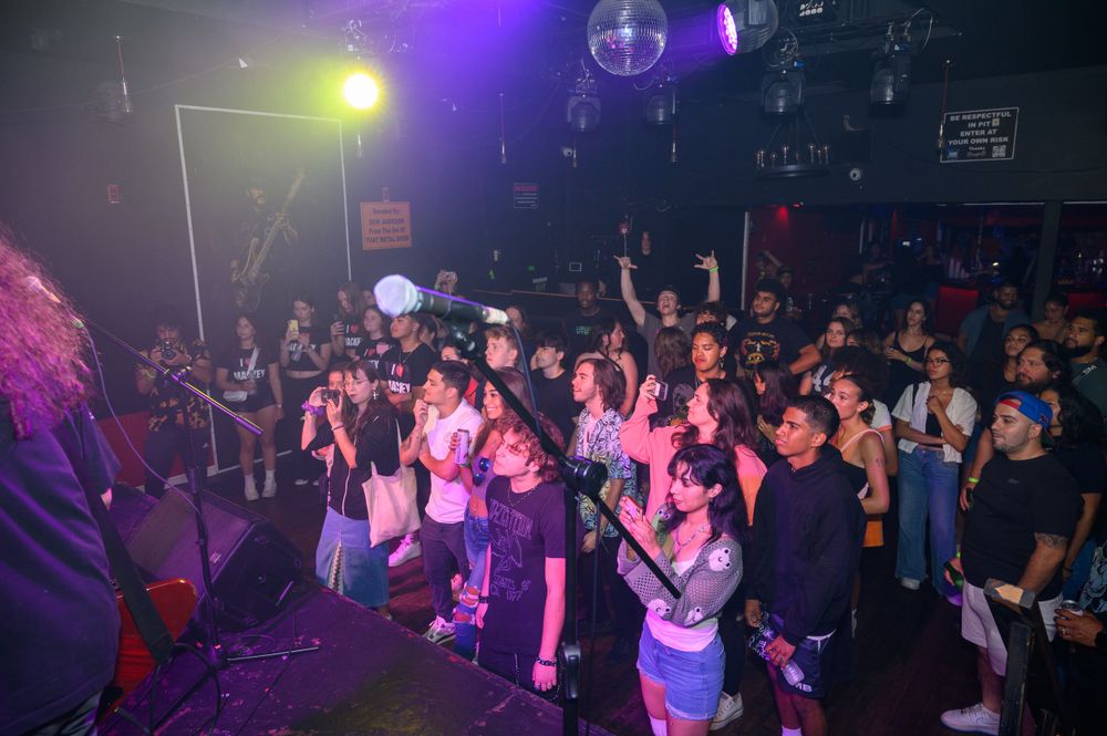 Concert crowd stands in front of stage with purple lighting and disco balls at a nightclub performance.