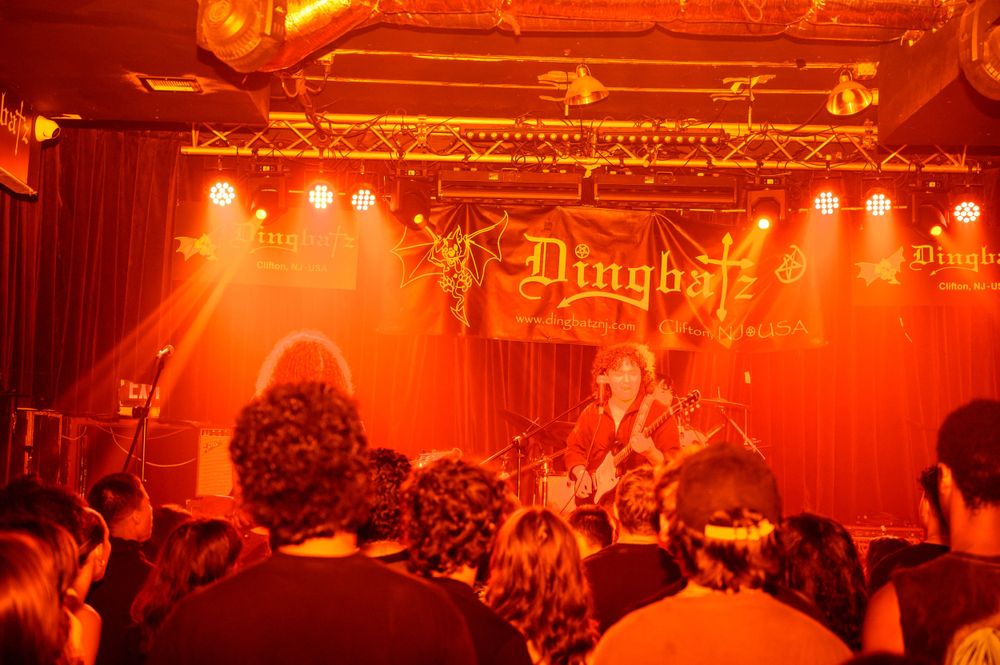 Concert crowd watches performers on stage bathed in dramatic red lighting at intimate music venue.